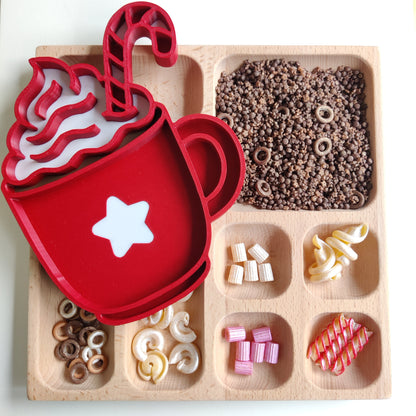 Hot chocolate inspired sensory play base., Red sensory play tray shaped like a coffee cup with a star on top, placed on a wooden tray with various small items.