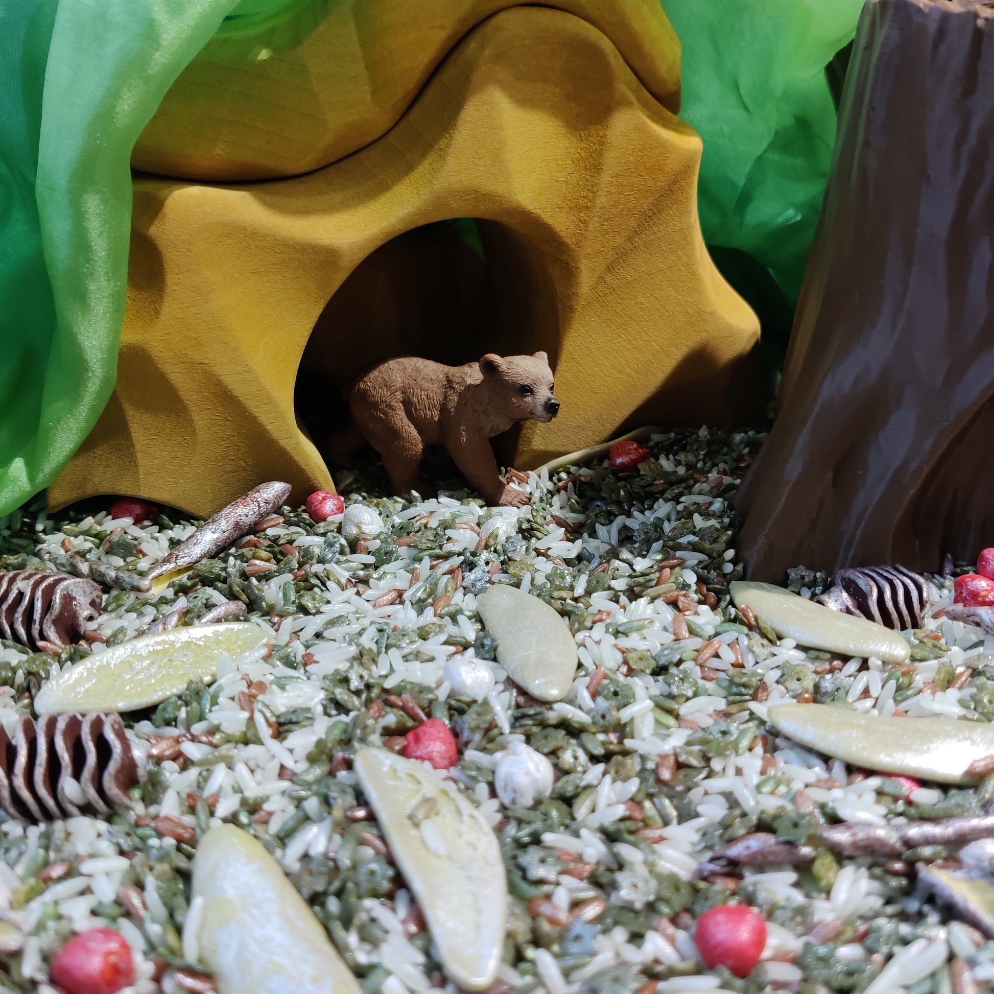 Small bear figurine peeking out from a yellow tunnel in a miniature forest setting with rocks and small plants.  Sensory play bases 